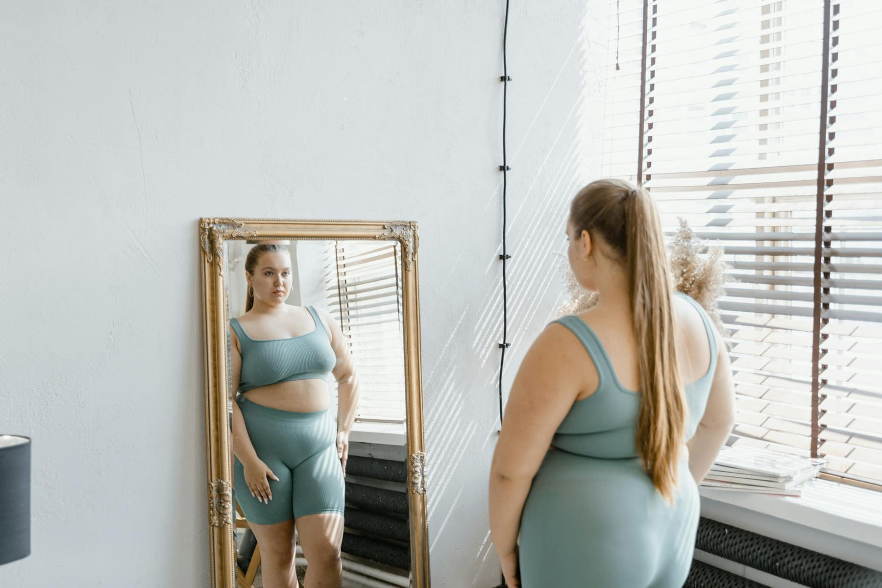 Woman in activewear embraces body positivity, standing before a mirror indoors. on Think Lean Live Strong